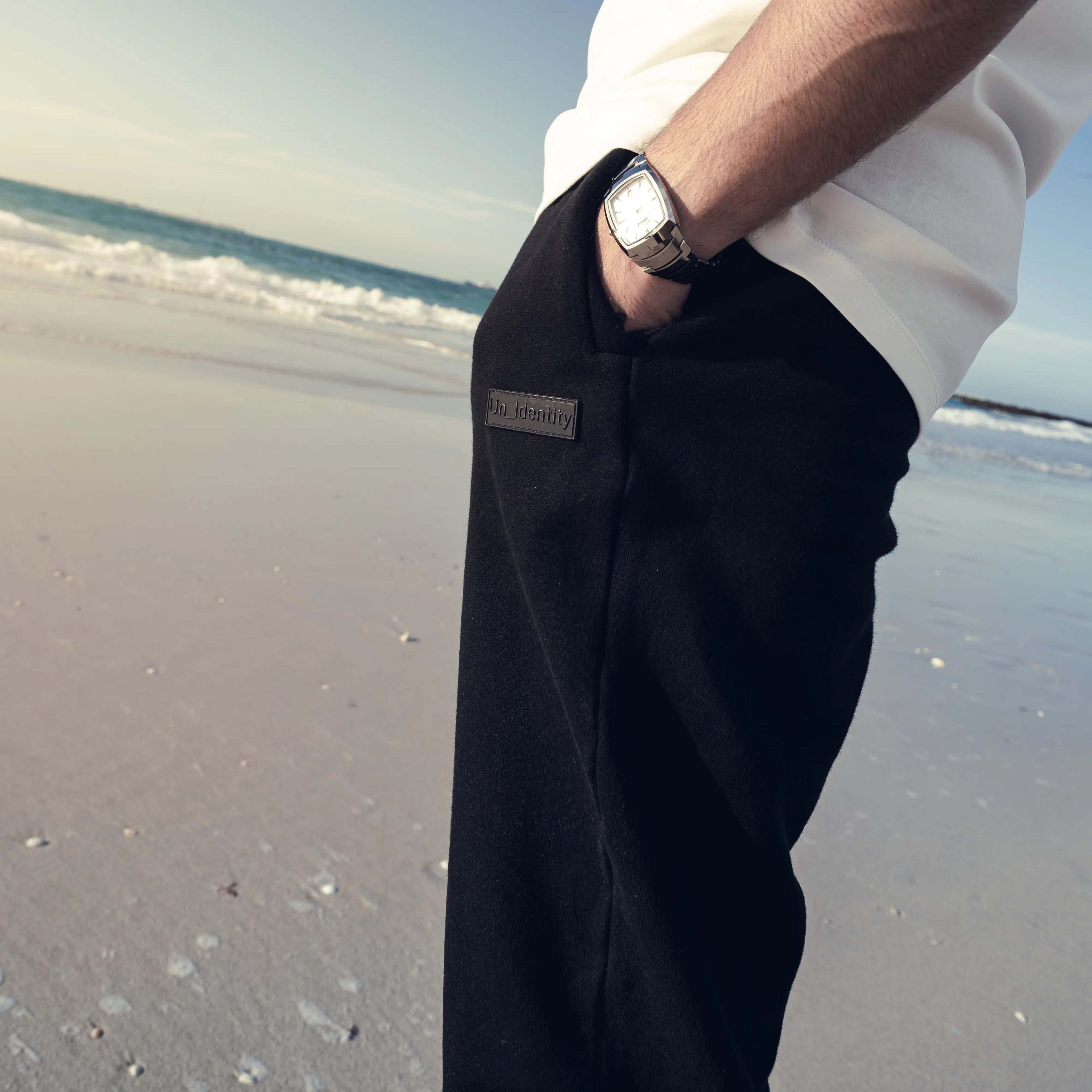 Person standing on a beach with ocean waves at the background wearing white shirt and black Unidentity pants