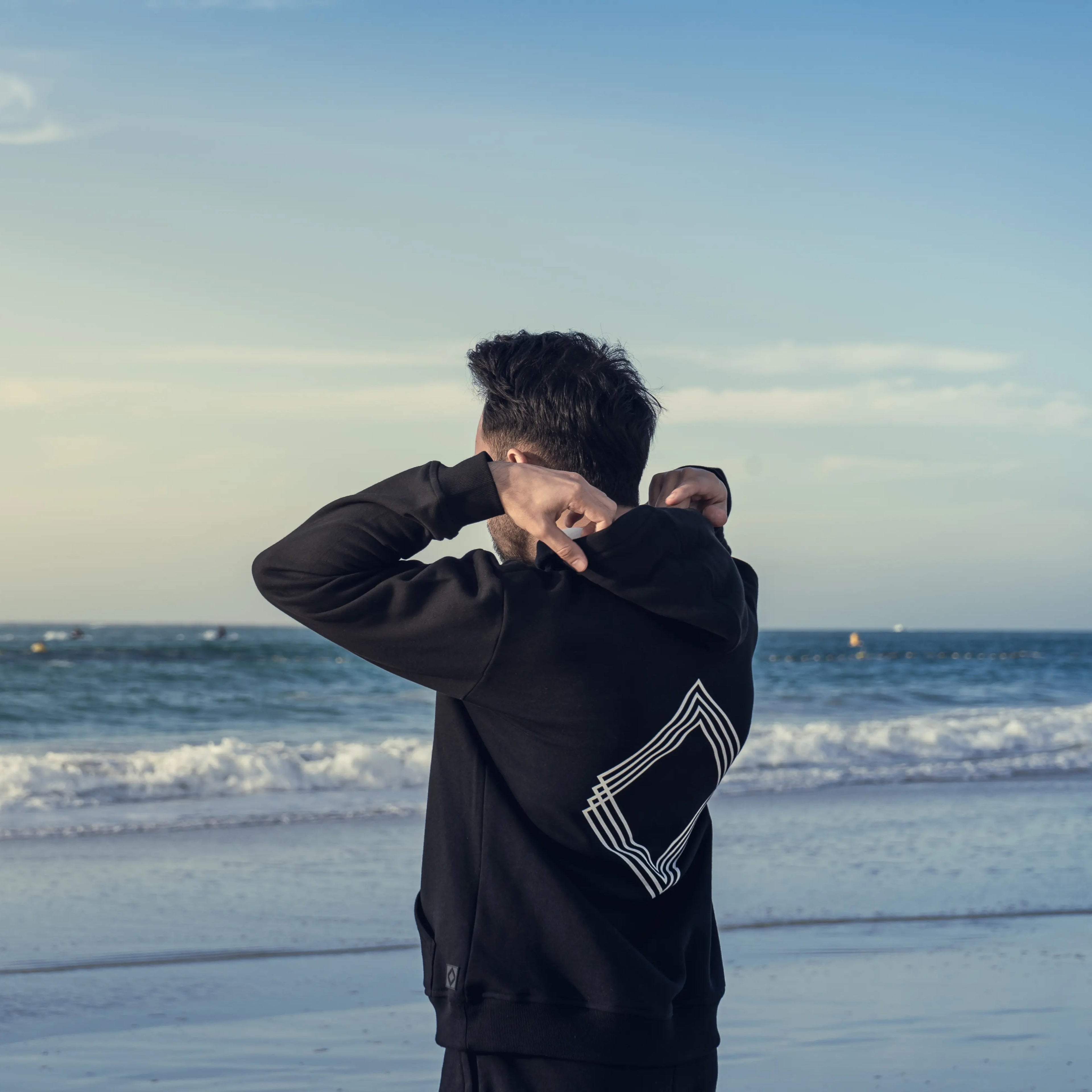 Man wearing black hoodie with white geometric design on a beach with ocean waves in the background