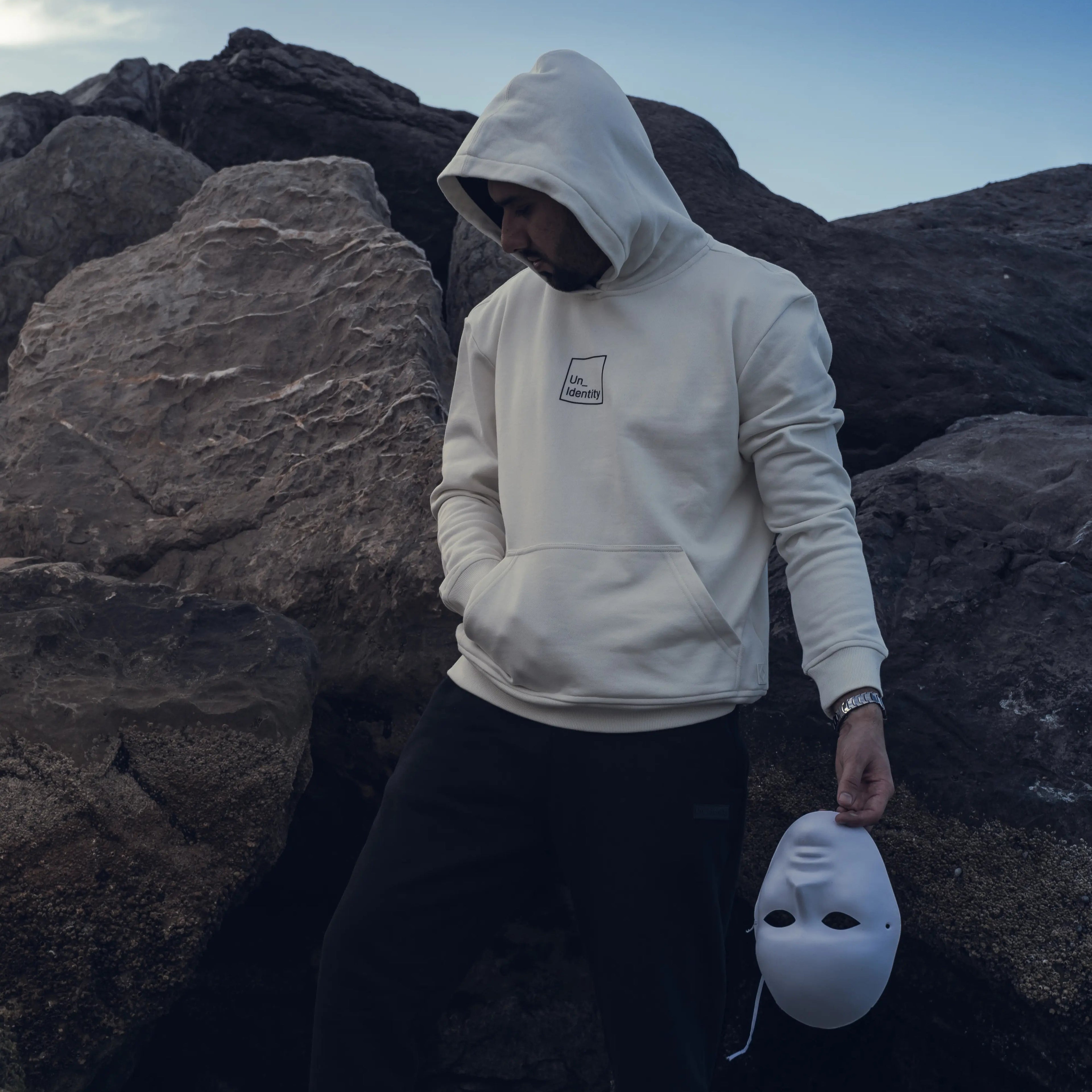 Person wearing white Unidentity hoodie and black pants standing on rocky terrain holding white mask.