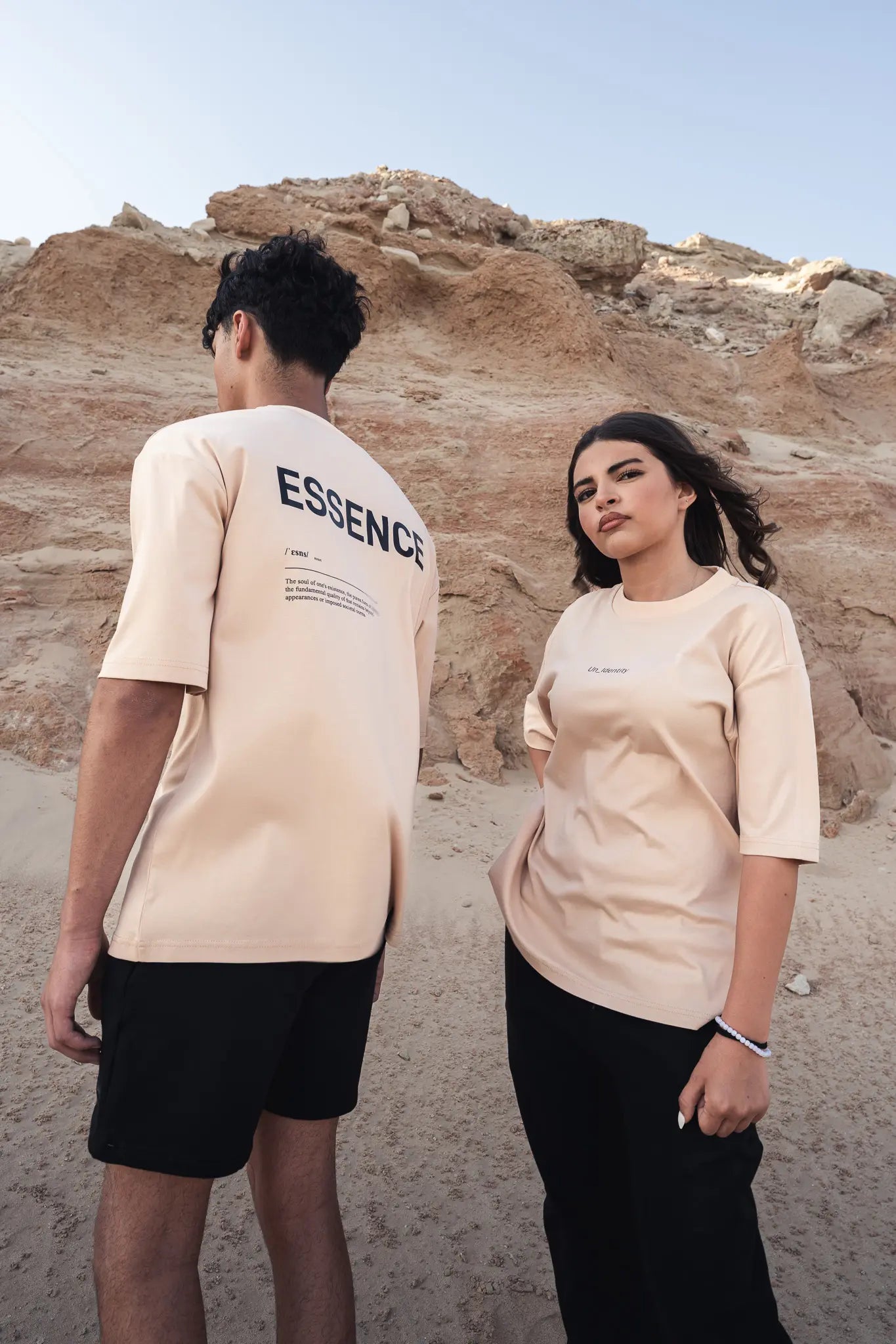 Two people wearing beige T-shirts with ESSENCE featuring in a desert setting. #color_beige