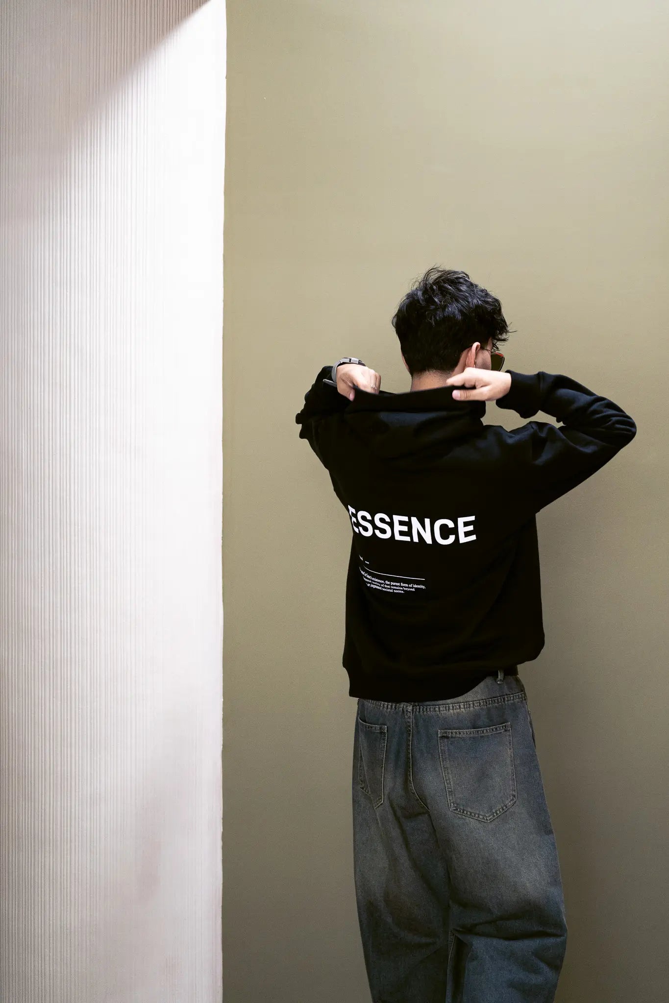 Person wearing black hoodie with white ESSENCE text on the back, standing against a plain wall