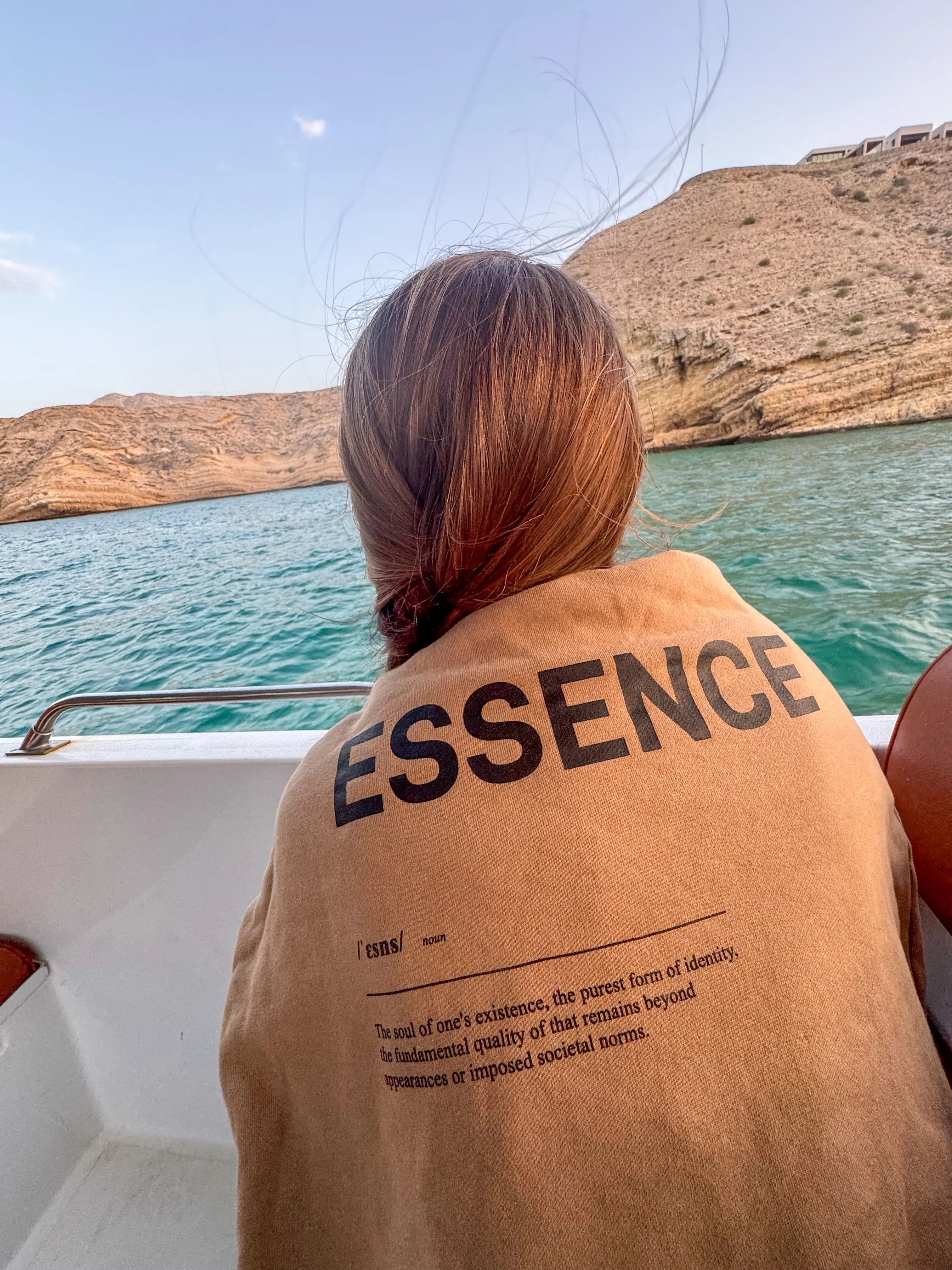 Person on a boat wearing brown garment with black ESSENCE text, overlooking a scenic ocean mountain view