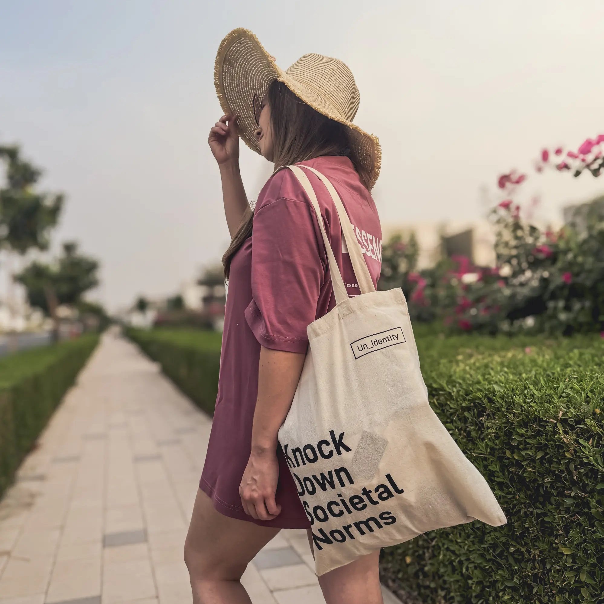 Woman holding Unidentity tote bag with text walking outdoors on a path with greenery and flowers