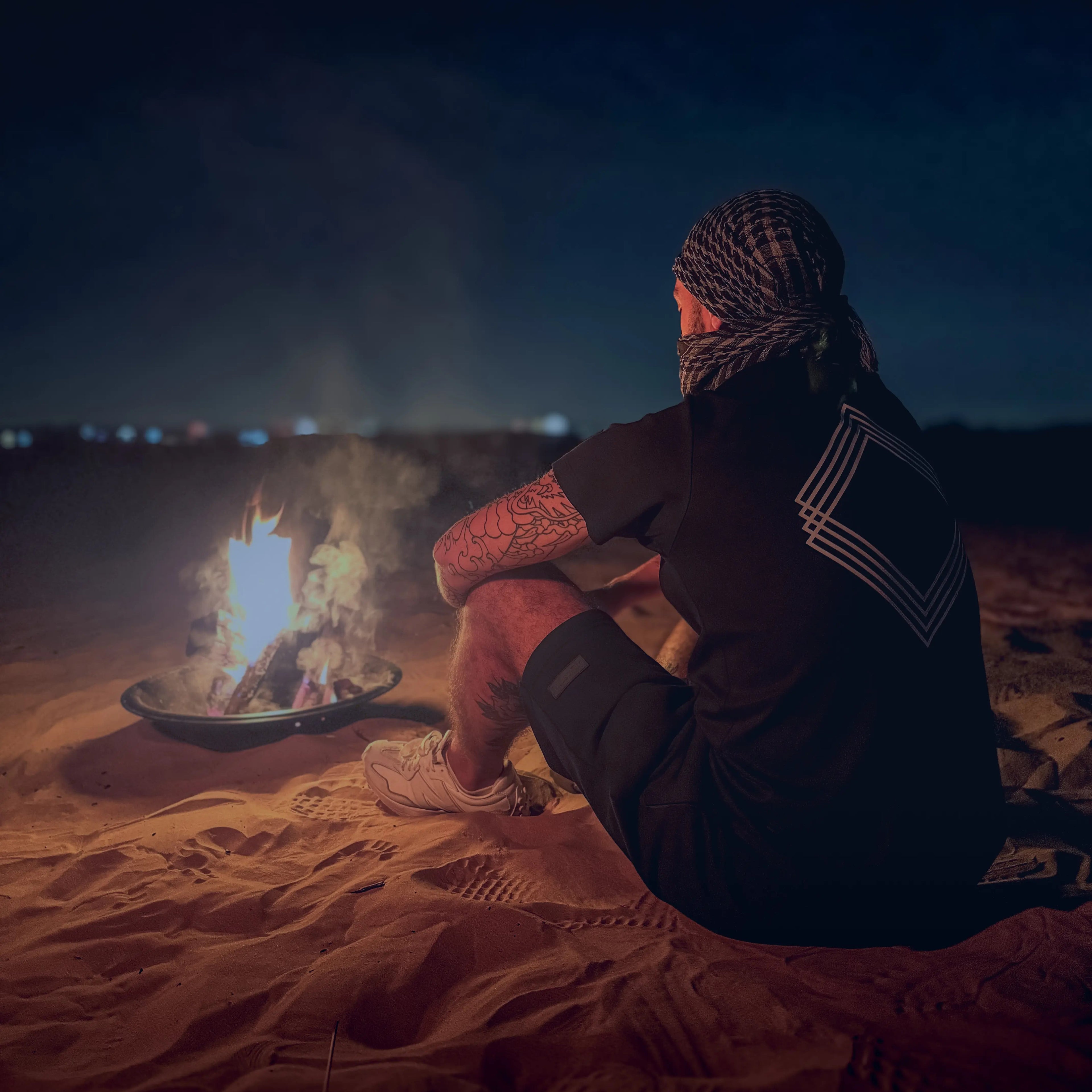 Man wearing Unidentity black shorts and tee sitting by a campfire in the desert at night