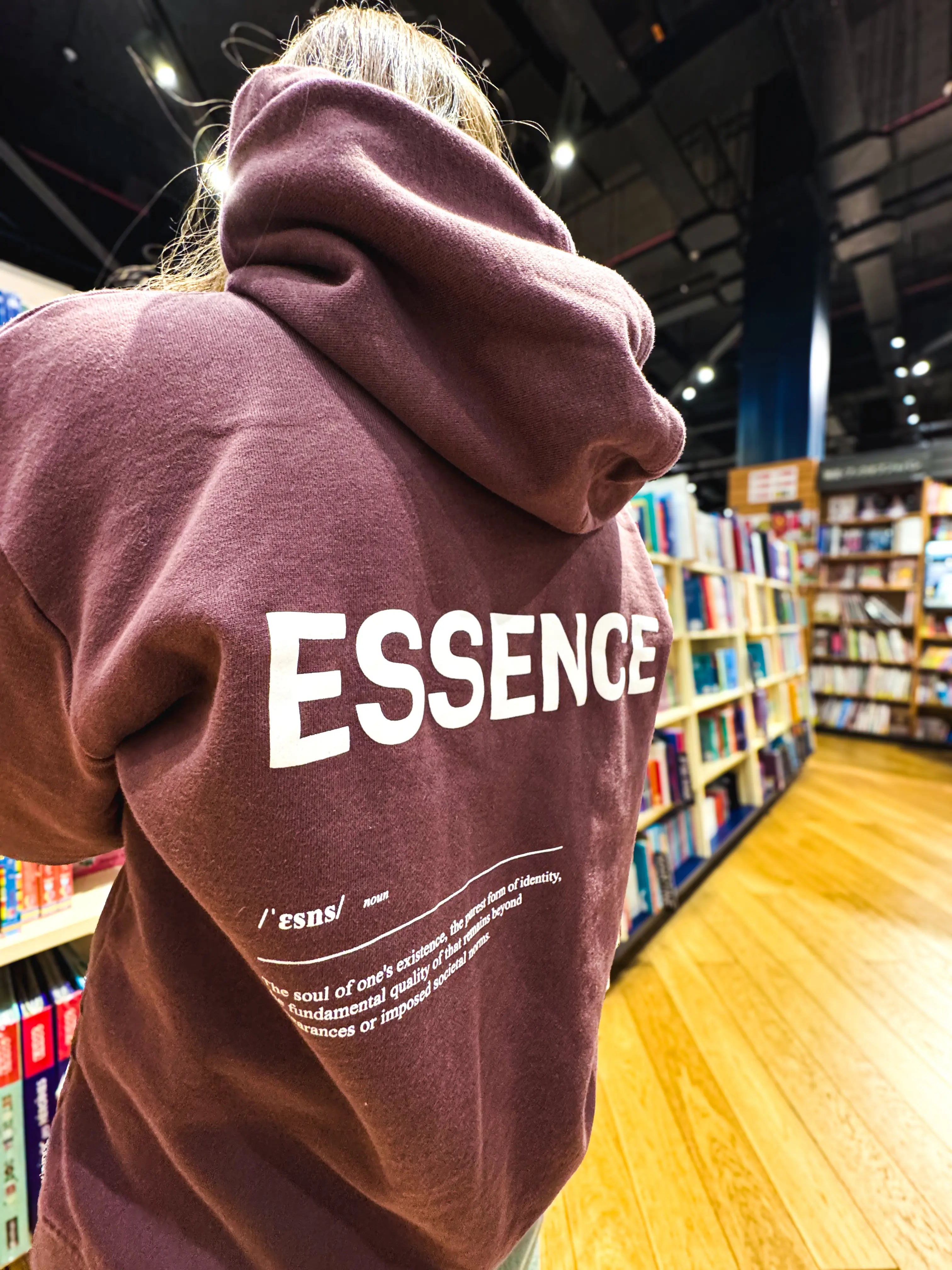 Person wearing Unidentity plum hoodie with ESSENCE text in a bookstore. #color_plum