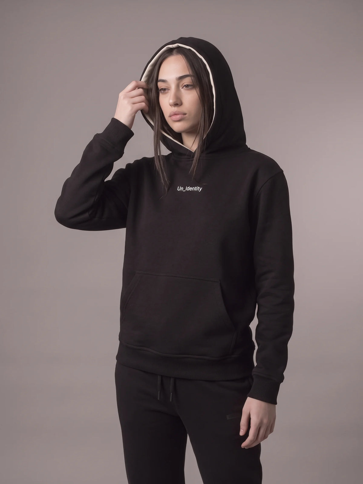 Black organic cotton hoodie, Unidentity Gem Hoodie, front view worn by woman with hood up and white Un_Identity wordmark, 390gsm #color_black