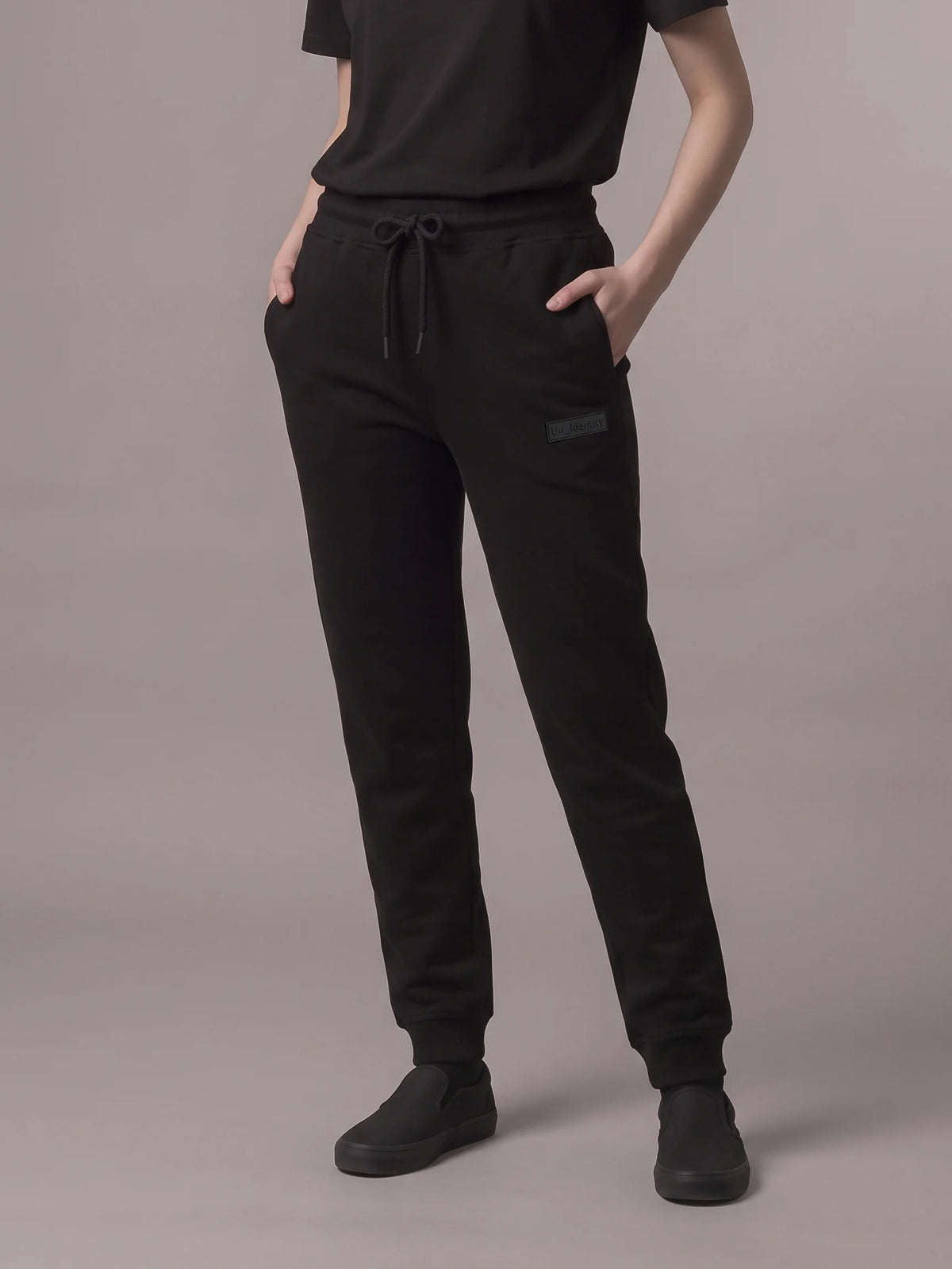 A model wearing black jogger sweatpants with a drawstring waist, side pockets, and ribbed cuffs, paired with a black t-shirt and black slip-on shoes, against a solid gray studio background. #color_black