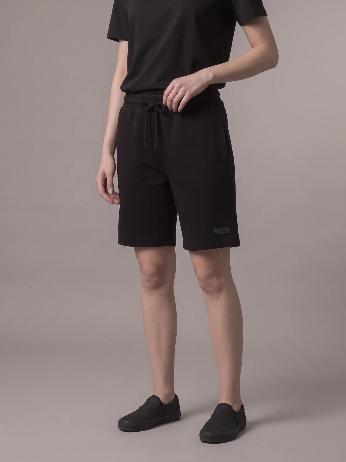 A model wearing black drawstring shorts, a black t-shirt, and black slip-on shoes, standing against a solid taupe background. #color_black