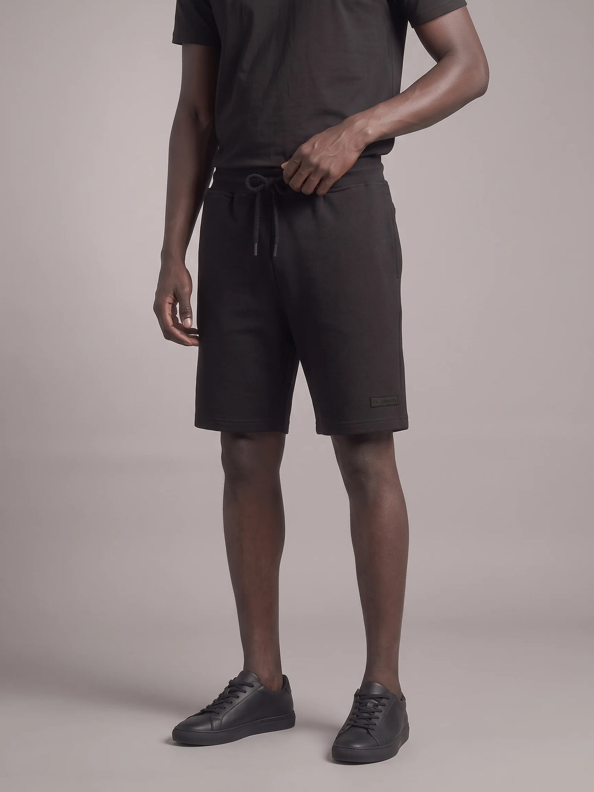 A model wearing dark charcoal gray drawstring shorts, a matching dark t-shirt, and black sneakers, standing against a neutral studio backdrop. #color_black