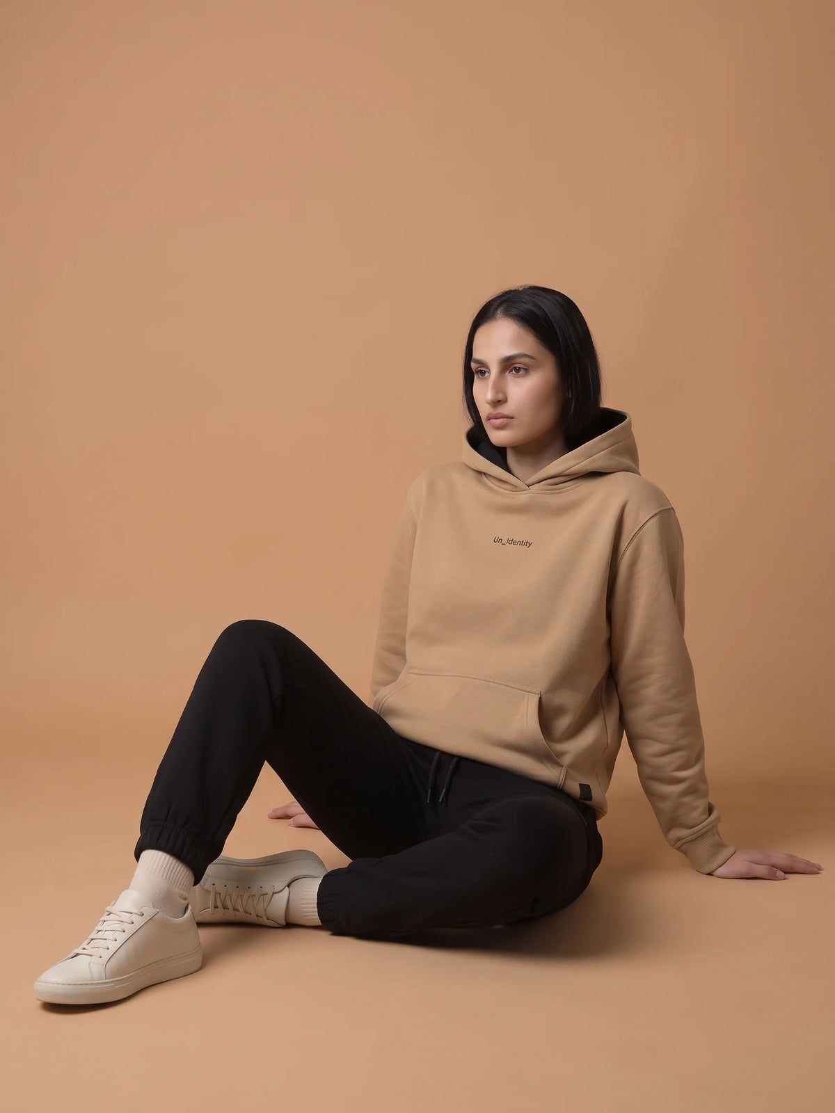 Beige regular fit hoodie, Unidentity Soul Hoodie, side front view worn by woman seated on the floor with black Core joggers, sustainable streetwear #color_beige