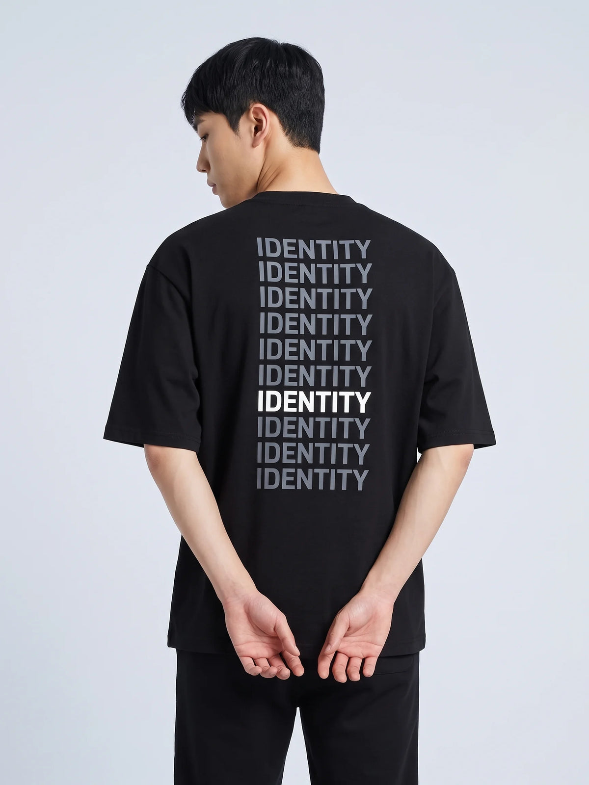Black oversized t-shirt, Unidentity Identity Tee, back close-up of the IDENTITY typographic print worn by man, hands clasped behind, streetwear #color_black