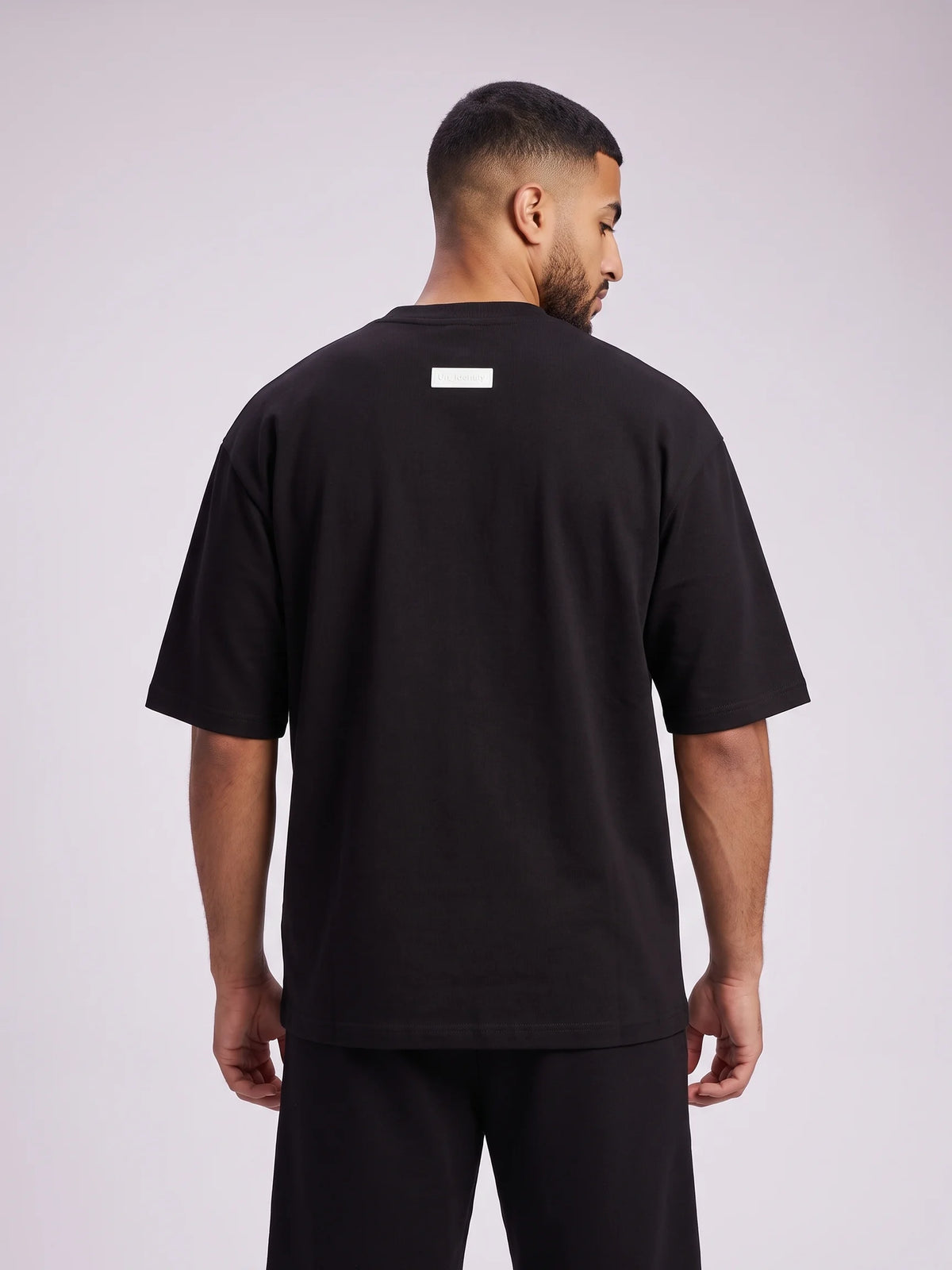 Black oversized t-shirt, Unidentity Foundation Oversized Tee, upper back view worn by man showing the drop shoulder and white 3D patch, streetwear #color_black