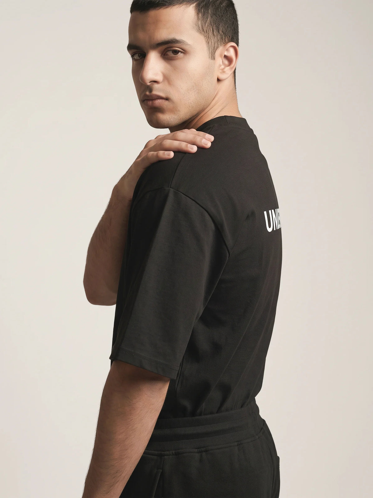 Black organic cotton t-shirt, Unidentity You Oversized Tee, side view of a man looking over the shoulder with the back logo just visible, 210gsm #color_black