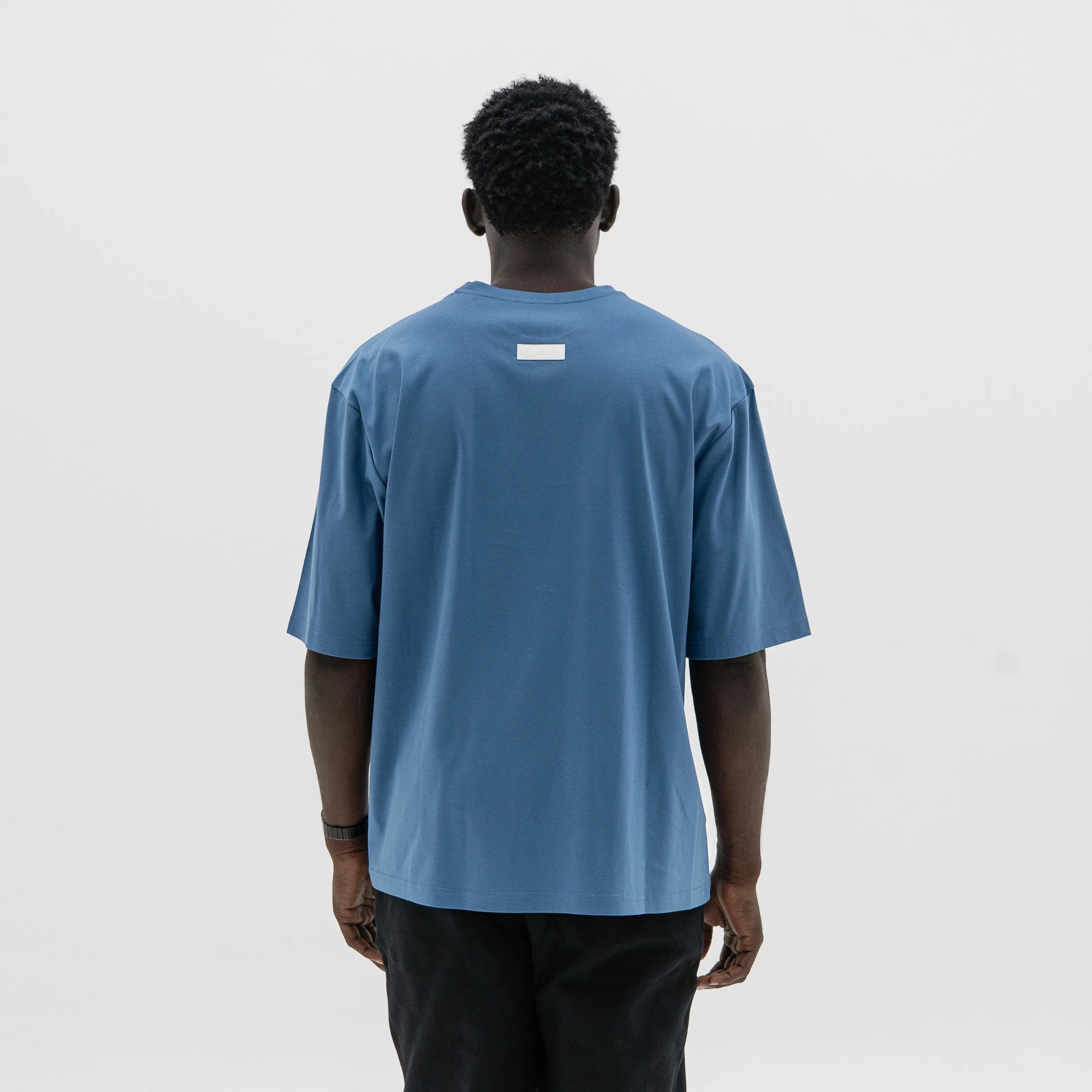 Person wearing a blue t-shirt with a small white logo on the back against a plain background. #color_blue