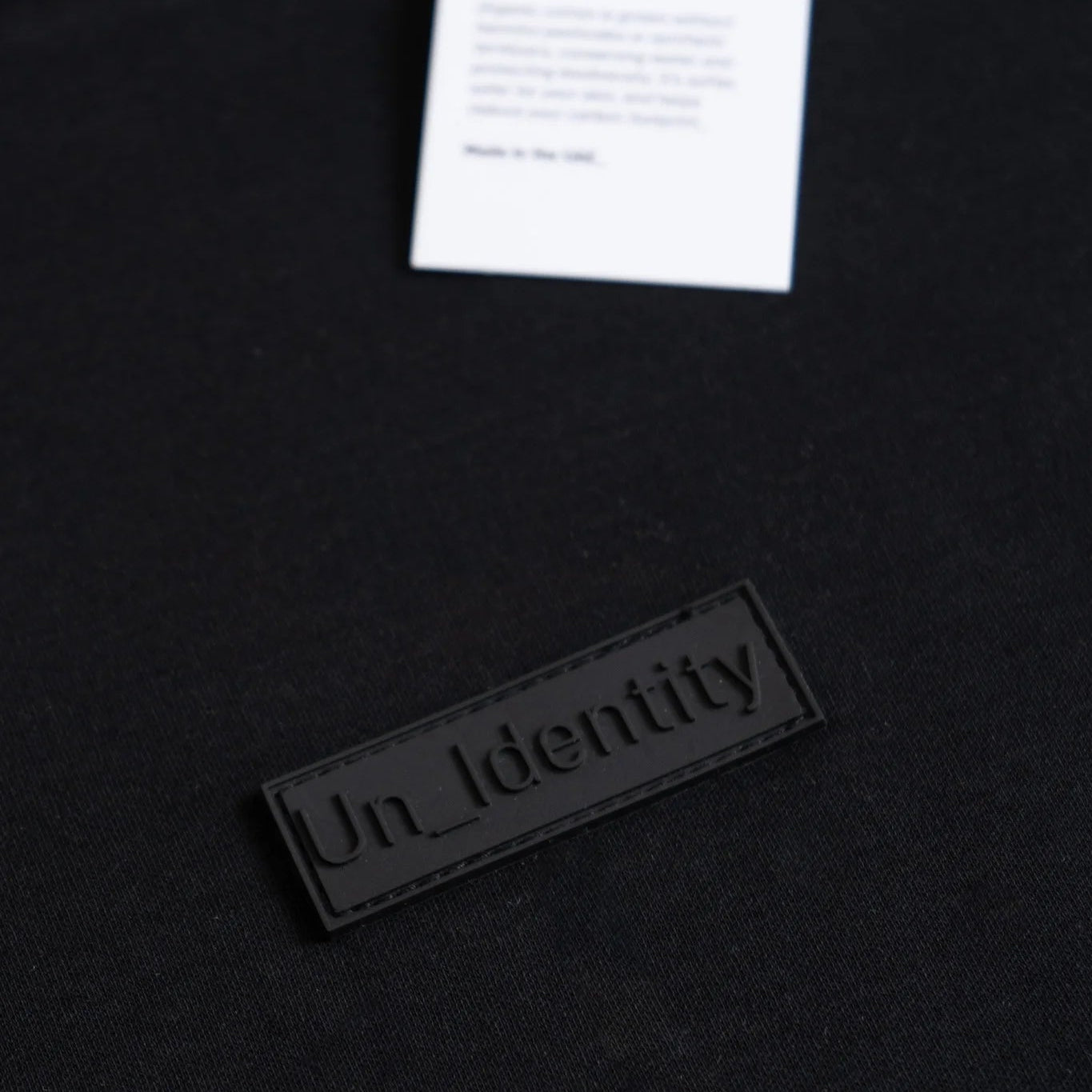 Black t-shirt with 'Un_Identity' label and white tag on a black background. #color_black