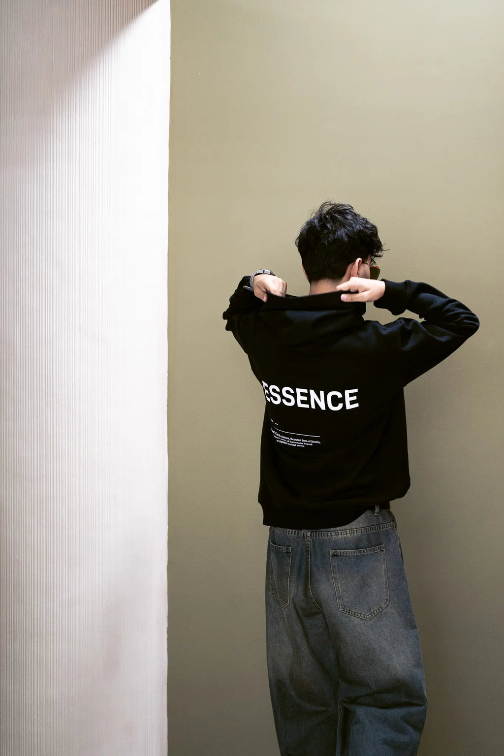 Person wearing black hoodie with white ESSENCE text on the back, standing against a plain wall