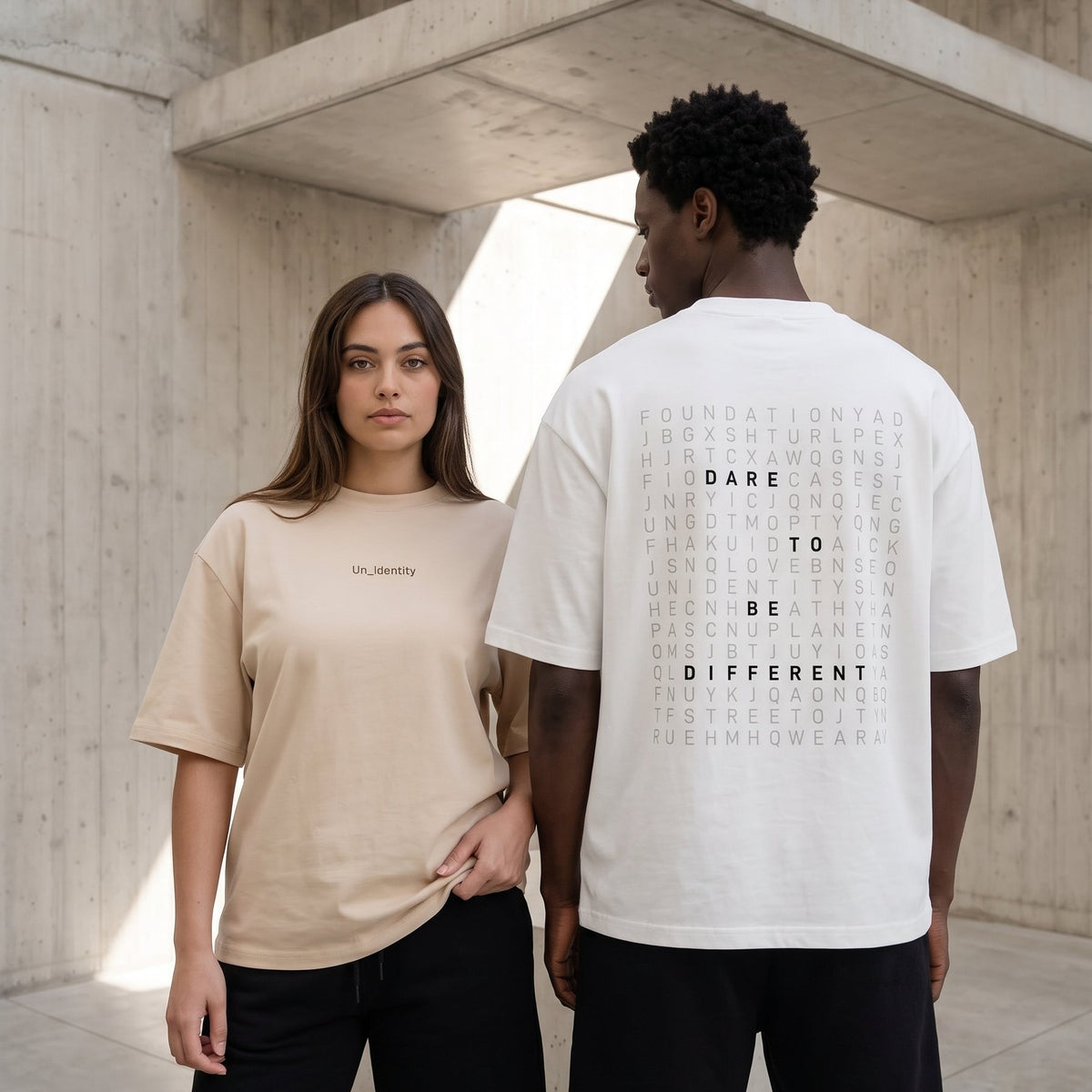 Two models wearing oversized graphic t-shirts in a concrete architectural setting. The model on the left wears a beige t-shirt with small black text on the chest, while the model on the right wears a white t-shirt from the back, showing a dense typographic design.