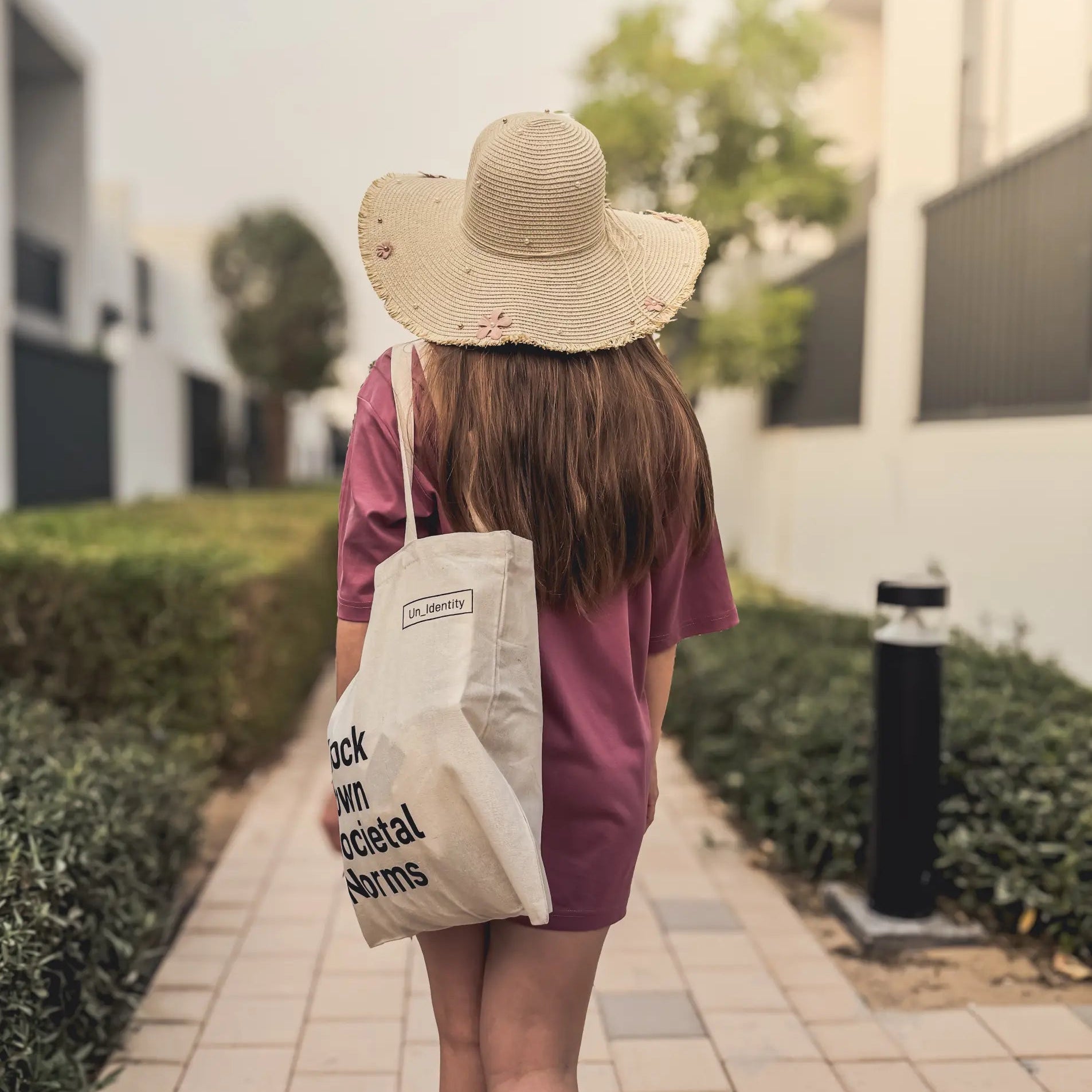 Girl wearing maroon T-shirt, straw hat and carrying Unidentity tote bag with text, walking on a sidewalk